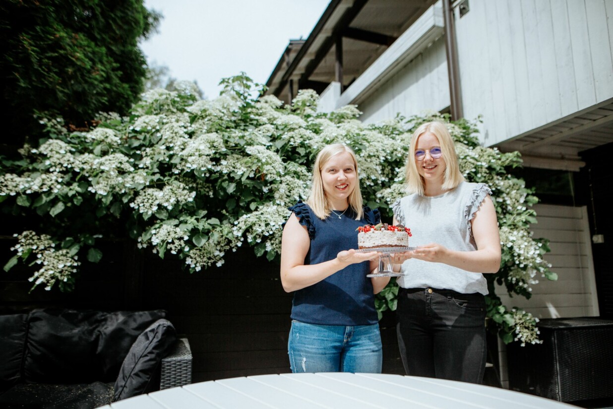 Kaksi tyttöä pitelee käsissään täytekakkua kukkivan pensaan edessä.