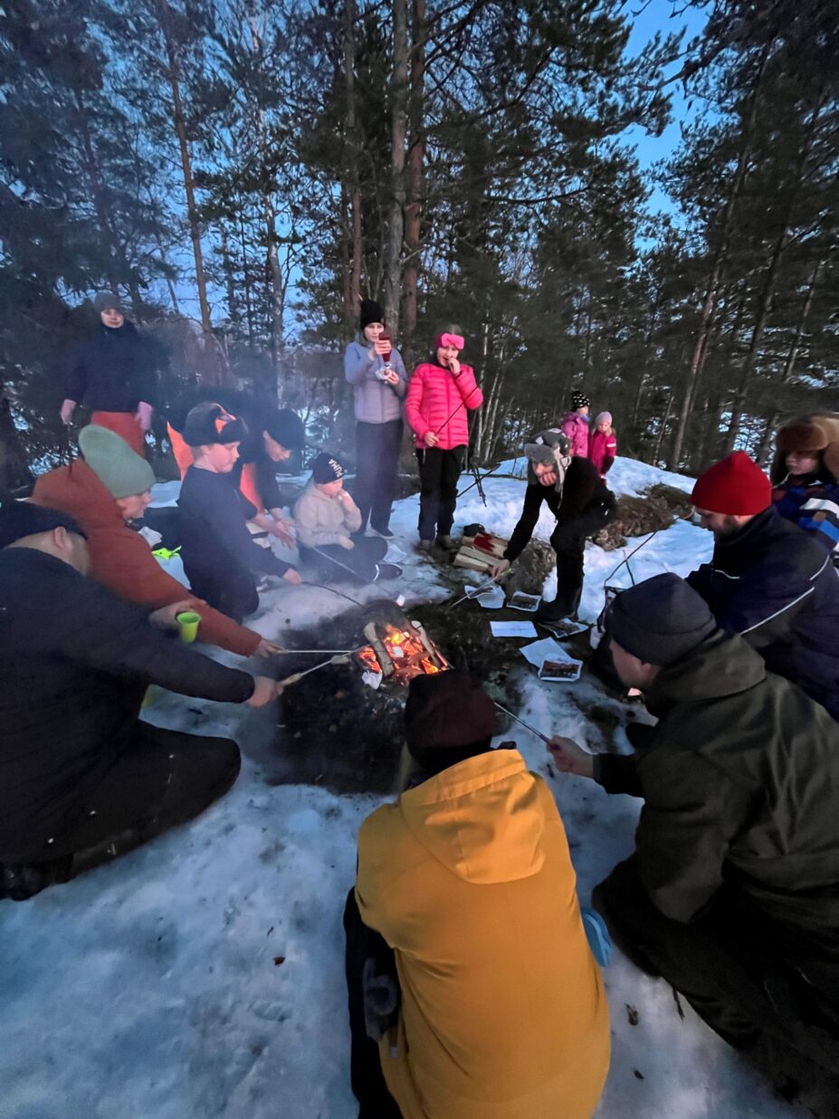 Joukko kerholaisia ja aikuisia kokoontuneena nuotion äärelle talvisessa metsässä.