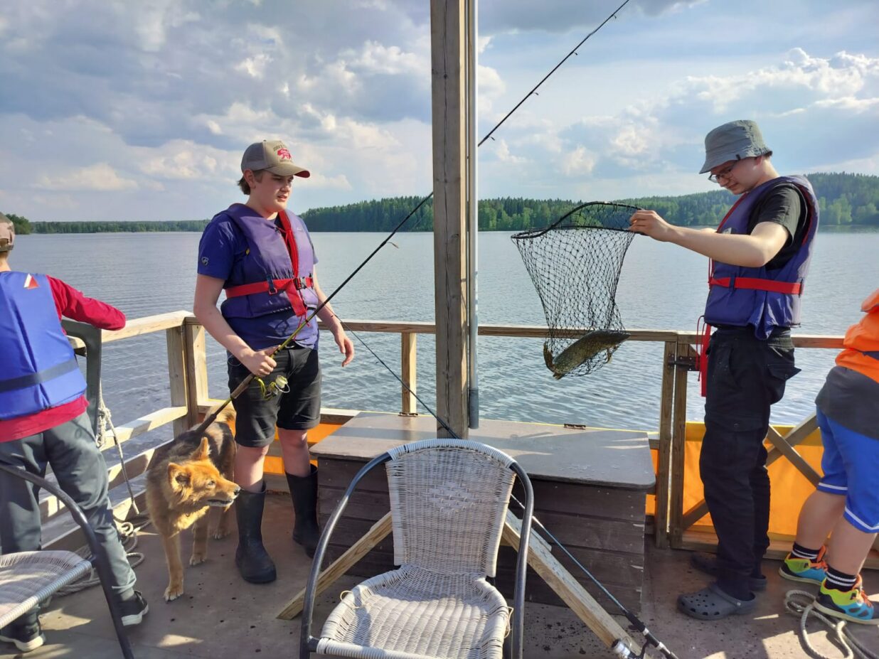 Kaksi nuorta ja koira veneessä. Toisella nuorista on kädessään haavi, jossa on kala.