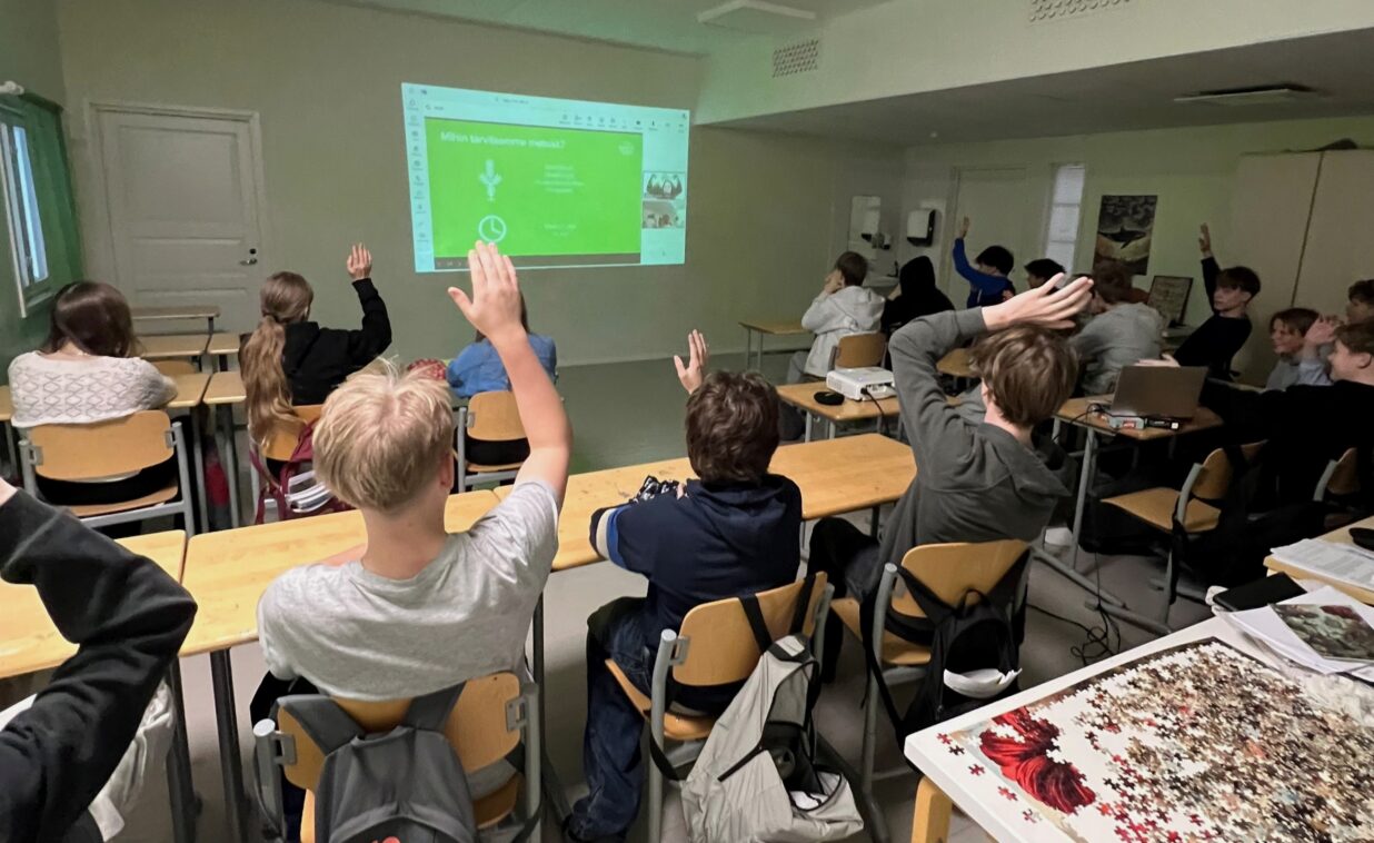 Kuvassa oppilaat seuraavat luokassa etäoppituntia. Osa oppilaista viittaa.