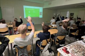 Kuvassa oppilaat seuraavat luokassa etäoppituntia. Osa oppilaista viittaa.